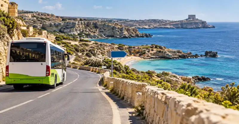 Autobús por una carretera costera con una playa de Malta al fondo.